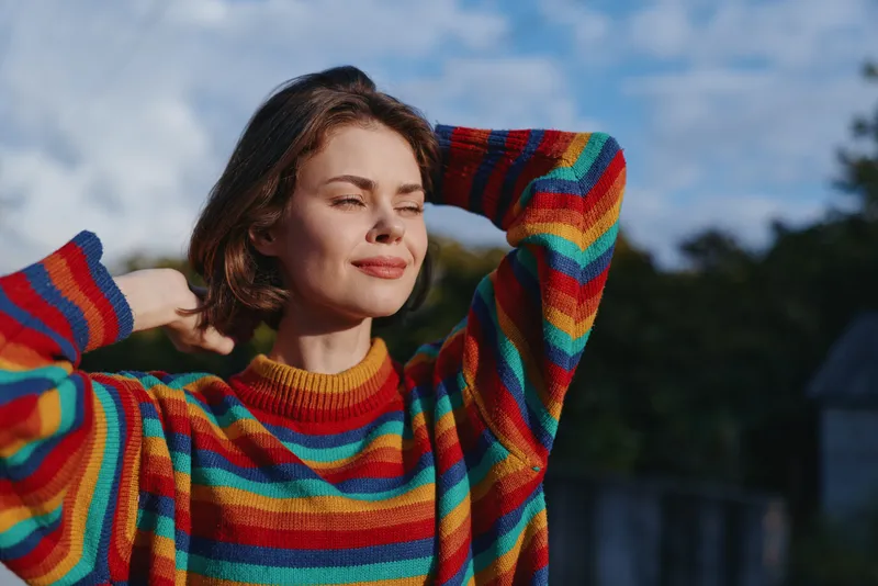 Woman in rainbow sweater smiling with eyes closed, young female model leaning back with hands behind head outdoors in sunlit park, relaxed cozy knit portrait for lifestyle fashion.