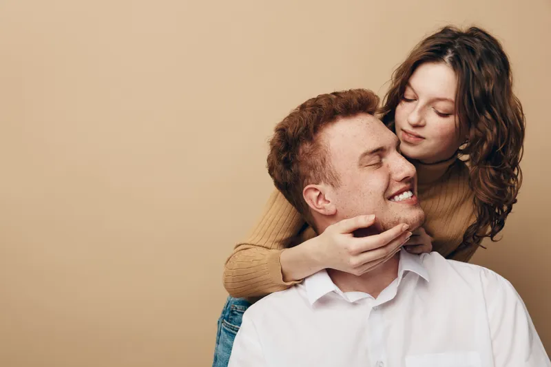 Young couple in casual clothes share a tender moment, woman embraces man from behind with gentle smile. Warm beige background creates intimate and loving atmosphere.
