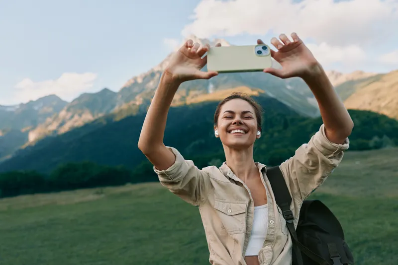 Casual woman outdoors takes a selfie in a mountainous landscape, smiling, enjoying nature, wearing a light shirt, backpack beside her, vibrant scenery