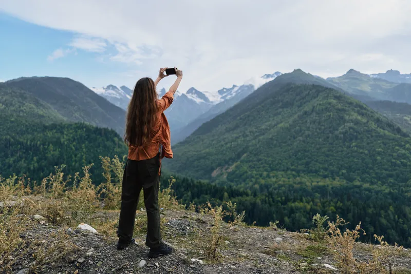 A person stands on a rocky overlook capturing a sweeping mountain landscape with a smartphone, snow capped peaks rise in the distance, clear sky, dramatic outdoor scene