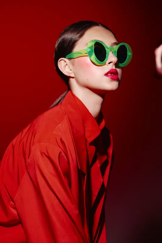 Young woman in oversized red clothing wears round green sunglasses. Her lips are vividly red and she poses dramatically against a solid crimson background. Fashion concept sharp lighting.