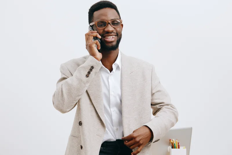 Smiling man with glasses talking on mobile phone while holding a tablet in a professional office environment, dressed in business casual, positive expression, bright background, lifestyle concept.