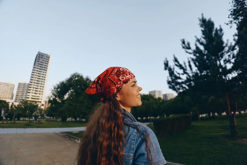 Lifestyle woman in modern boho-western denim street style, wearing a red bandana and smiling outdoors with urban buildings and greenery under warm film color tones.