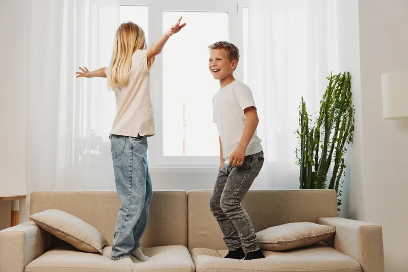 Happy children jump on sofa in bright living room, enjoying playful moments together. Concept of childhood joy, family fun, and active indoor entertainment with natural light and casual clothes.