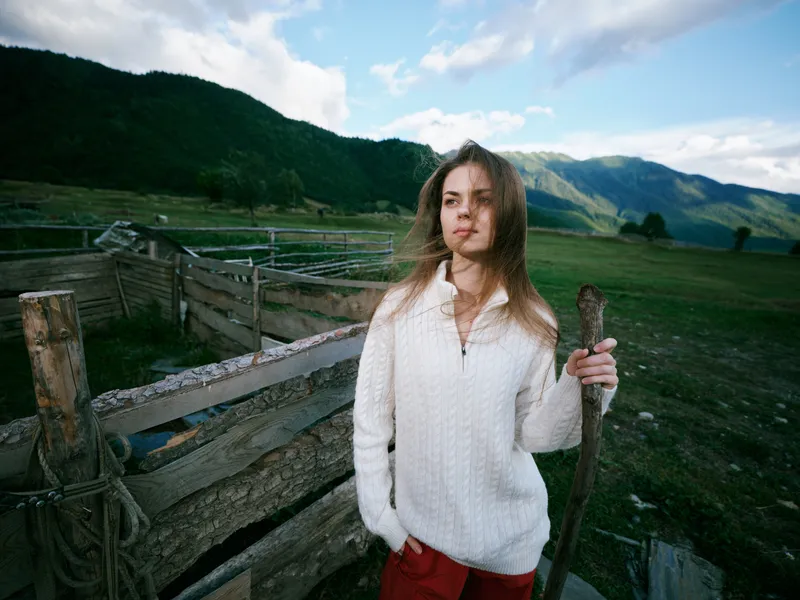 Countryside scene with a woman in a white sweater standing beside a wooden fence in a green valley, capturing a calm outdoor rural landscape and open sky with breeze and hills
