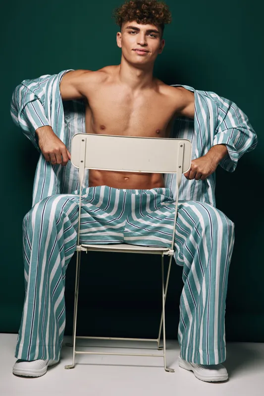 Young man with curly hair wearing striped pajamas sitting backwards on metal chair against dark background. Casual pose, relaxed mood and comfortable homewear fashion style.
