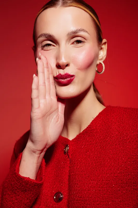 Close-up of a stylish woman with red lipstick and hoop earrings whispering with hand near mouth. Red background and red textured jacket create a bold, fashionable portrait concept.