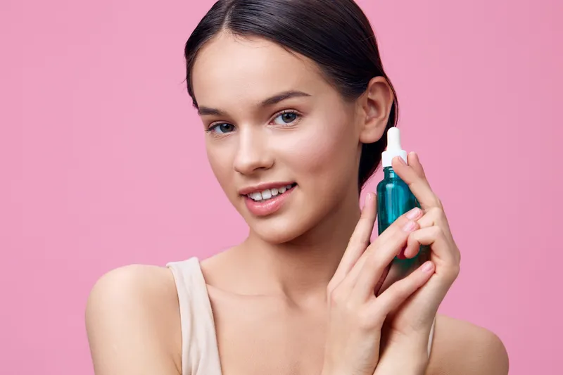 Young woman holding a skincare serum in front of a pink background, promoting beauty and self care with a healthy glow and smiling expression