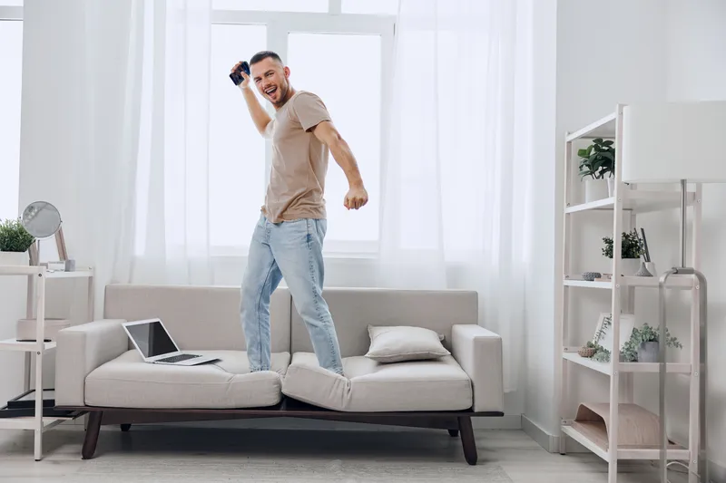 Energetic man celebrating success while standing on a couch, dressed casually in light clothing, with a joyful expression that reflects a modern lifestyle.