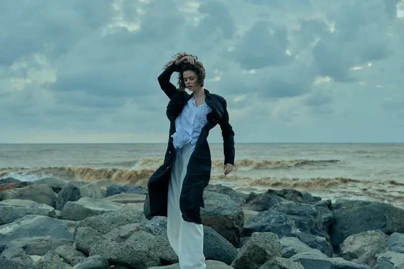 Woman in a long coat braves a windy shoreline, stepping on jagged rocks by the sea, captured in motion with stormy sky and churning water for a dramatic outdoor fashion mood.