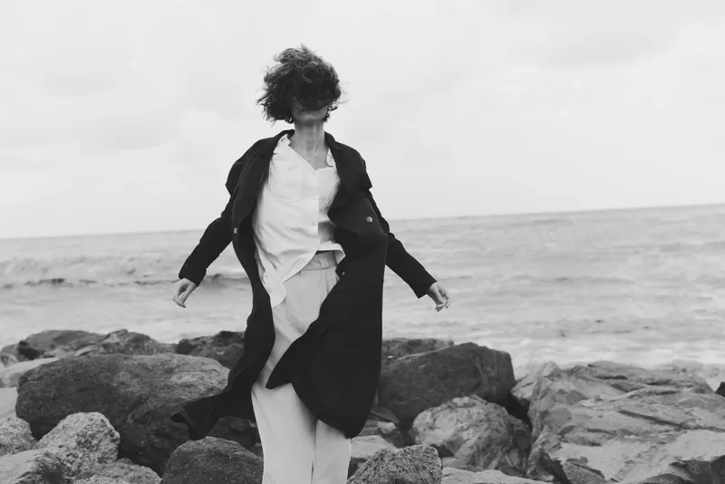 Woman on rocky shoreline, wind sweeping coat as she moves beside the sea in a moody monochrome scene with rugged textures and open horizon