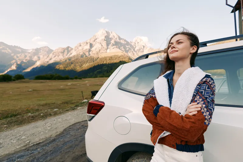 Woman by car and mountains on a roadtrip, travel portrait showing relaxed smile and confident pose near SUV in nature. Cozy sweater, sunlight over meadow and scenic outdoor adventure.
