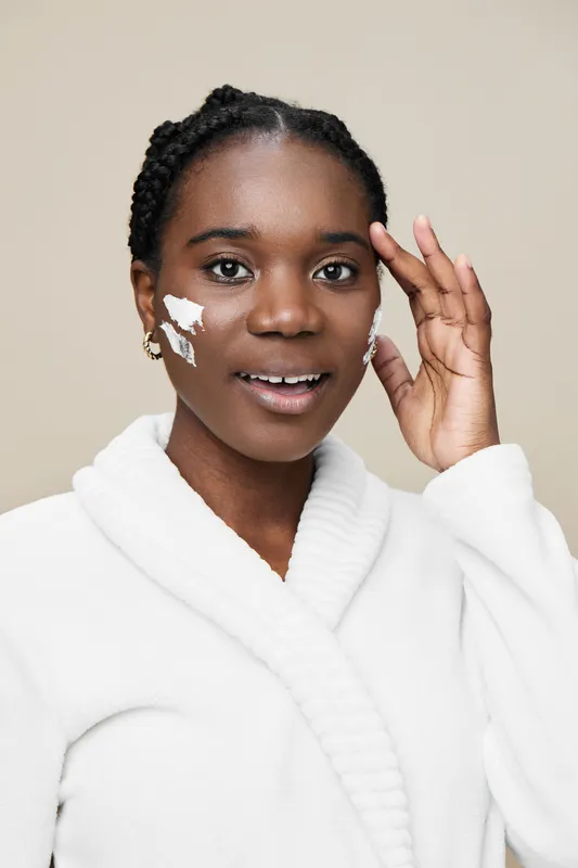 A joyful young woman of African descent applying skincare with a smile, wearing a cozy white robe, showcasing healthy skin and self care
