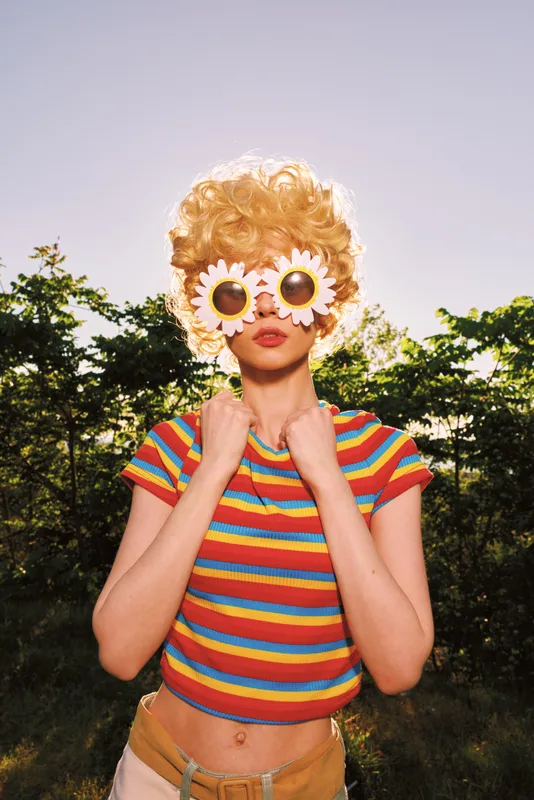 Smiling girl portrait outdoors in fashion, colorful whimsical playful nature summer rainbow scene, with daisies over eyes and a striped top, creating a bright warm mood