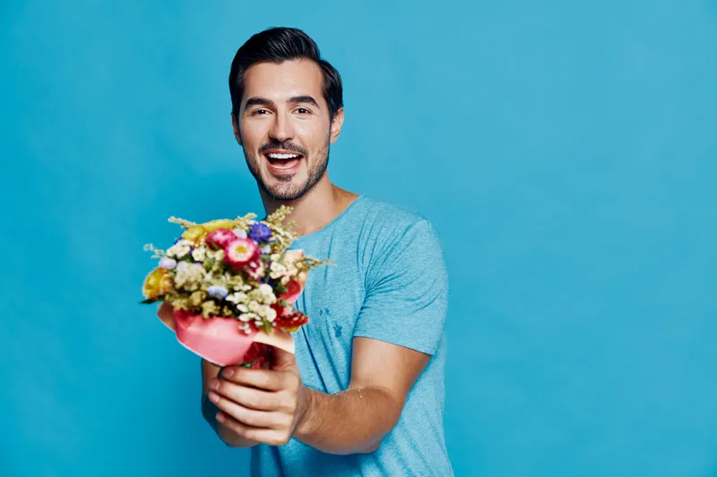 Happy smiling man holding a vibrant bouquet of flowers, wearing a casual blue t-shirt, with a joyful expression against a bright blue background. Floral concept.