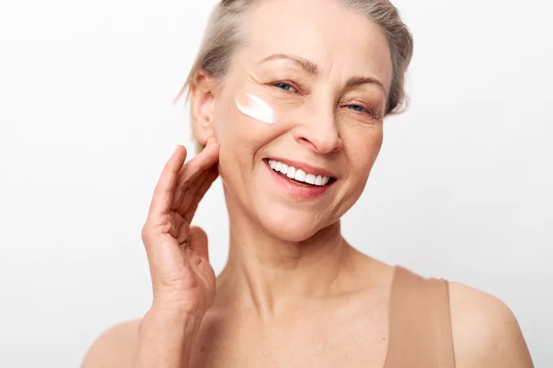 Smiling mature woman with clear skin applies face cream on cheek. Close-up portrait on white background. Concept for skincare, anti aging, beauty routine, and healthy lifestyle.