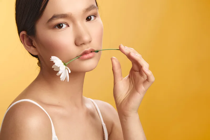 asian woman delicately holds a white flower near her lips against a warm yellow backdrop, creating a graceful beauty shot with soft lighting and calm, refined presence.