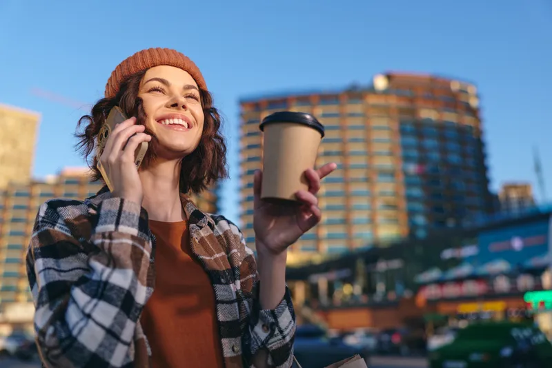 woman coffee phone smile city urban golden candid authenticity mindful living emotional storytelling Young woman chats on phone holding takeaway coffee in warm golden hour glow, joyful urban street