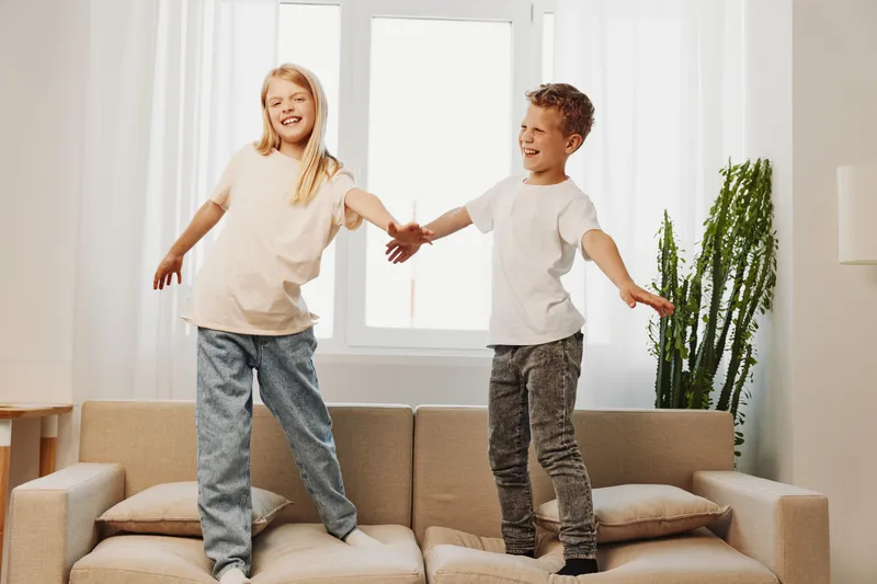 Two happy children, boy and girl, jump on a beige sofa in a bright living room with natural light. Concept of childhood joy, playful moments, sibling fun, and home happiness.
