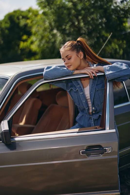 Young woman in denim outfit leaning on car door, looking relaxed outdoors with greenery, evoking a sense of freedom and adventure.