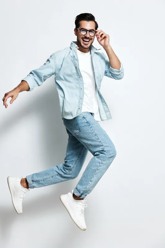 Full body smiling happy cheerful young man wearing denim shirt and glasses jumps joyfully while looking at the camera against a light solid background. People lifestyle concept