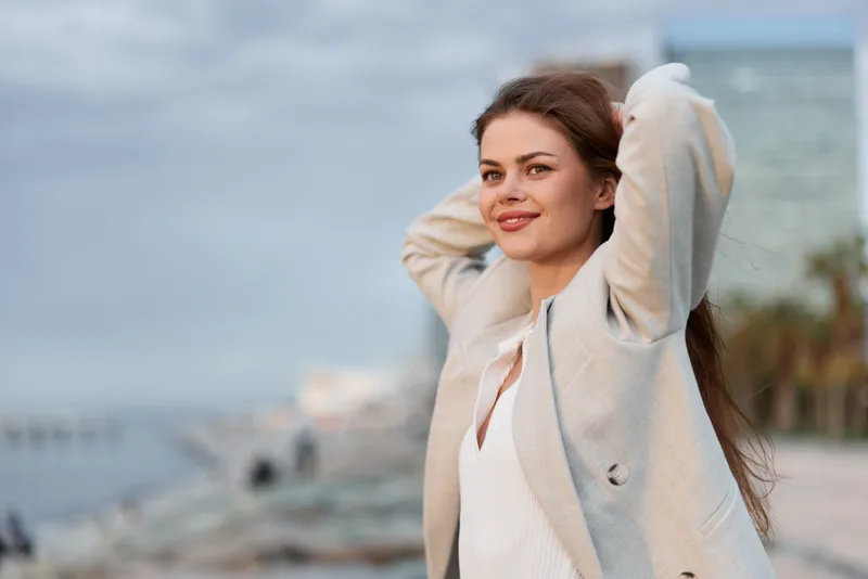 Confident young woman in beige coat outdoors smiling with relaxed pose by waterfront, autumn fashion, natural light, urban background, positive emotion