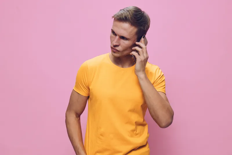 Casual young man with short brown hair wearing bright yellow t-shirt talking on mobile phone isolated on light purple background. People lifestyle concept, communication, casual style, cheerful mood, 