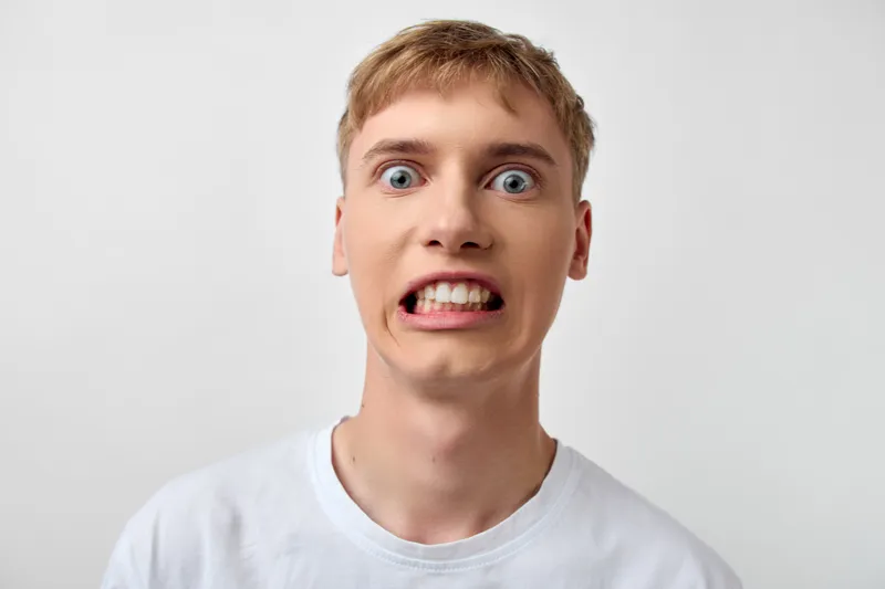 Surprised man with wide eyes and gritted teeth in a white t-shirt, closeup facial expression showing astonishment, dramatic emotion in a studio setting