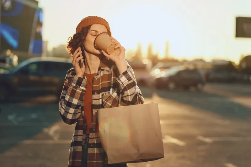 Young woman drinking coffee from a takeaway cup while on the phone with a shopping bag in urban sunlight; candid lifestyle scene with authenticity, golden hour glow and emotional storytelling.
