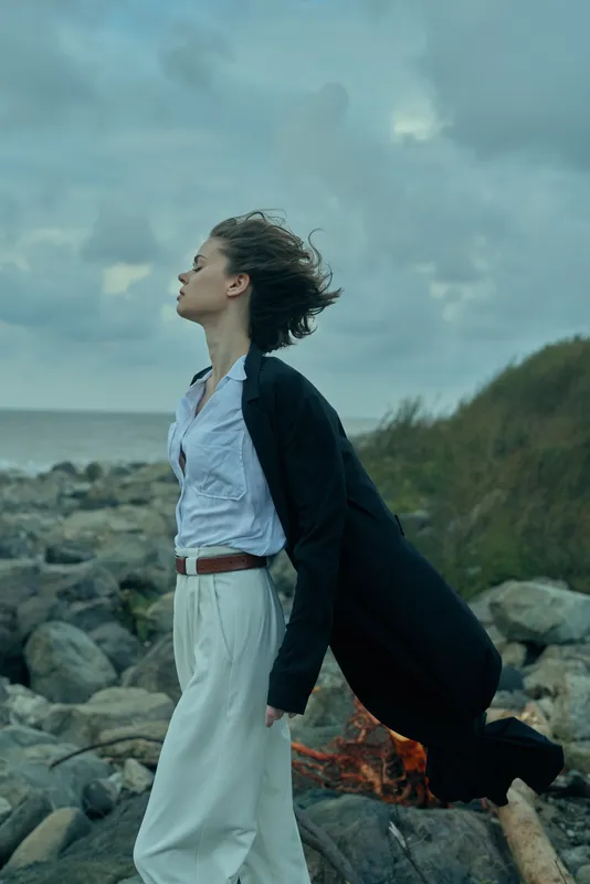 Woman walking along a rocky coast as wind lifts her coat, sea breeze and stormy clouds create a dramatic outdoor moment of freedom and resilience