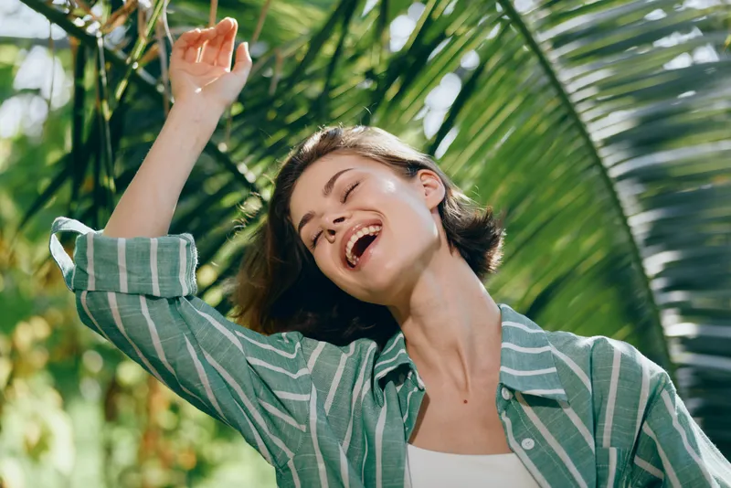 Joyful young woman laughing in bright sunlight, arms raised, wearing a striped green jacket, with lush palm leaves framing a carefree outdoor moment.