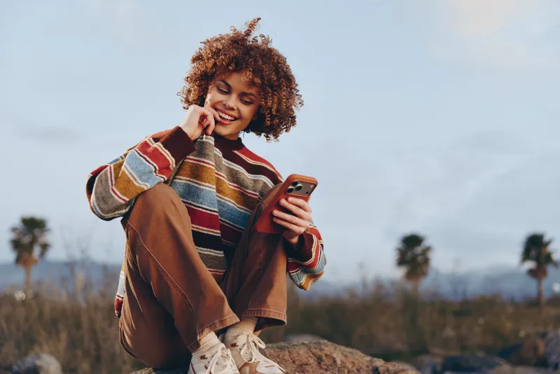 With a smile, woman outdoors in a field holds a smartphone and touches her ear, wearing a striped jacket, curly hair in wind, lifestyle moment in golden sunset, open landscape, calm mood