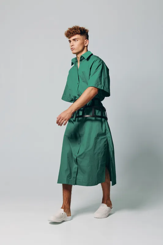 Young man wearing green oversized shirt and matching skirt with white sneakers standing on gray background in studio, fashion model posing with casual streetwear style.