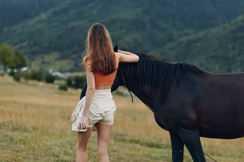 A young woman stands beside a horse in a rural field, lightly touching the animal's neck. She wears an orange top and light shorts, calm, connected, and serene in nature.