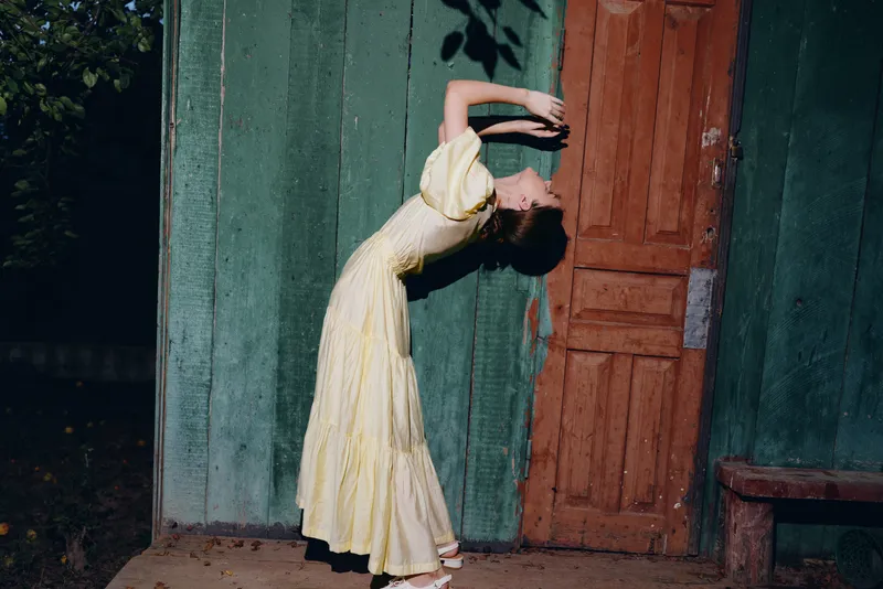 woman dancer in pale yellow dress arches backward beside a weathered green wall and wooden door, capturing graceful movement and expressive pose in an outdoor artistic scene