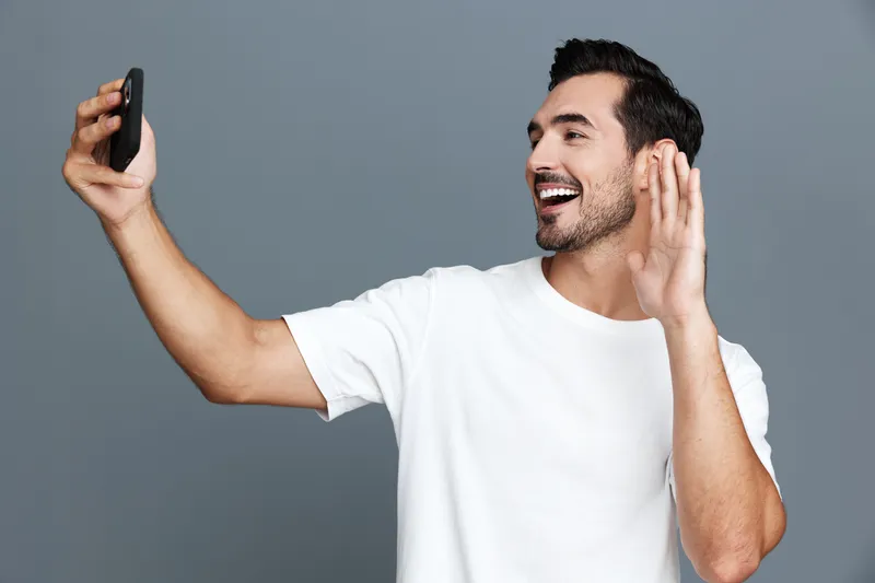 Happy cheerful young man waving hand while making a video call using mobile phone, isolated on plain gray background. People lifestyle concept.