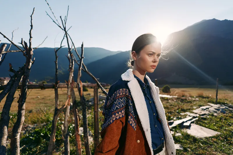 Woman portrait in countryside mountains with warm sunlight wearing a wool coat by a rustic fence, thoughtful young traveler captured in golden backlight and scenic valley landscape.