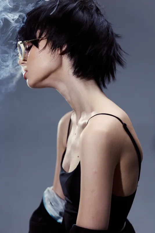 Profile of a stylish woman with short black hair exhaling smoke in a dark outfit, modern and moody concept