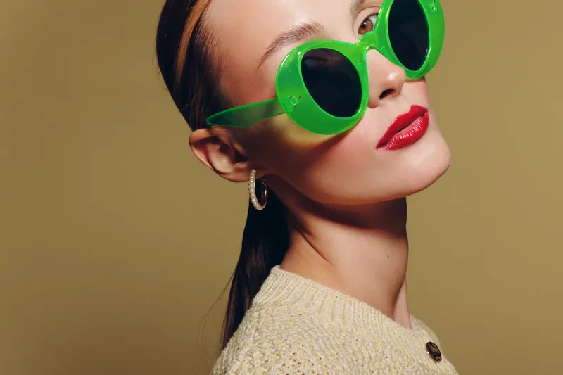 Stylish woman wearing large green sunglasses and red lipstick poses against beige background. Fashion portrait with trendy accessories and confident expression in studio lighting.