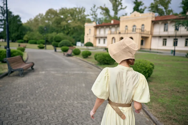 Elegant woman in a light dress and woven hat walks along a courtyard path toward a historic estate, creating a serene vintage ambiance and timeless travel atmosphere with soft sunlight