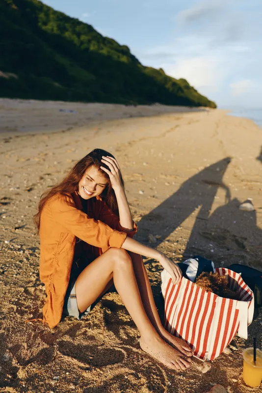 Woman on beach sand, sunlight summer travel relax, tote striped bag nearby, coast sea background, smiling seated pose, orange sweater, calm outdoor lifestyle, warm day