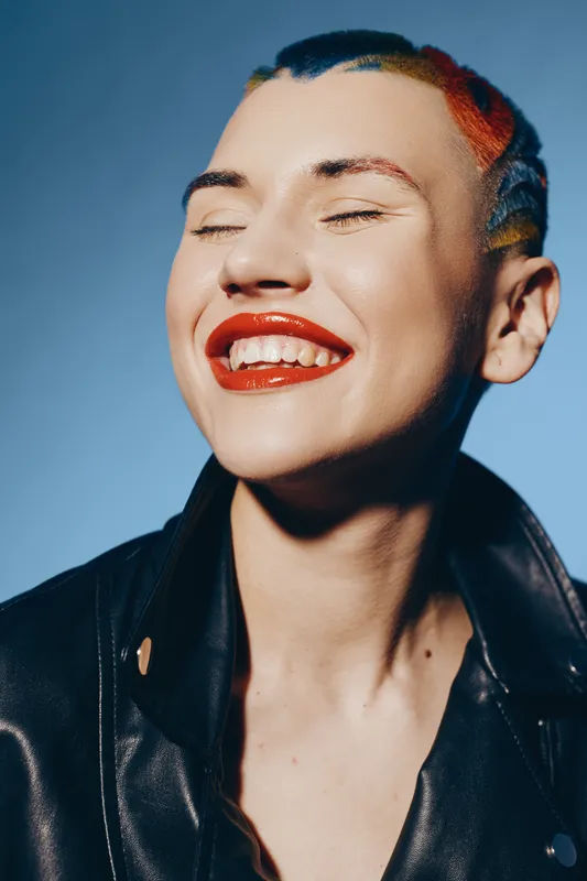 Smiling woman with colorful dyed hair and bright red lipstick wearing a black leather jacket, expressing joy and confidence against a plain blue background.