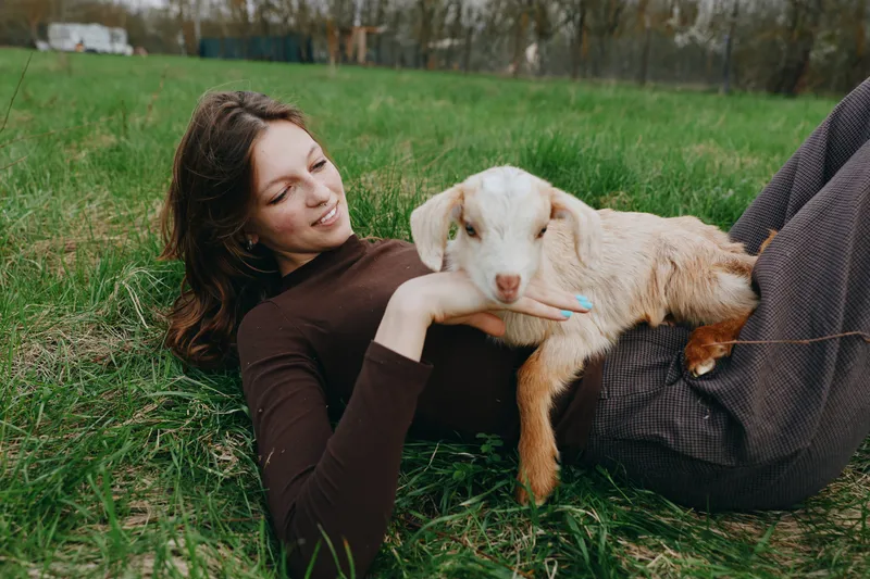Young woman lying on grass smiling while holding a baby goat in a casual outfit enjoying a peaceful moment outdoors in a lifestyle setting during spring.