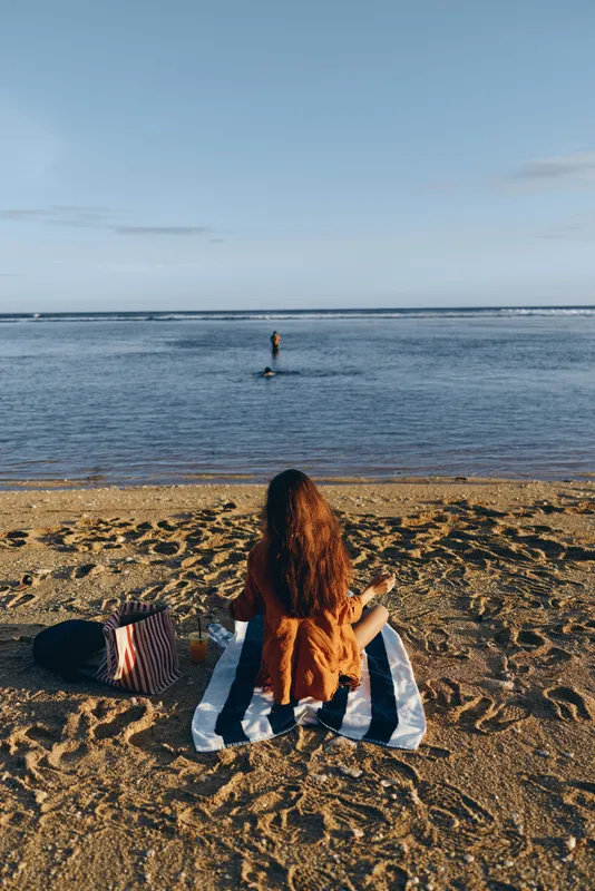 Young woman with long brown hair sits on a striped towel on the sandy beach watching a person standing in the calm sea under a clear blue sky.