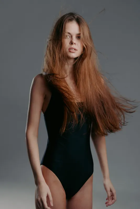 Young woman with long flowing hair wearing black bodysuit standing against neutral background