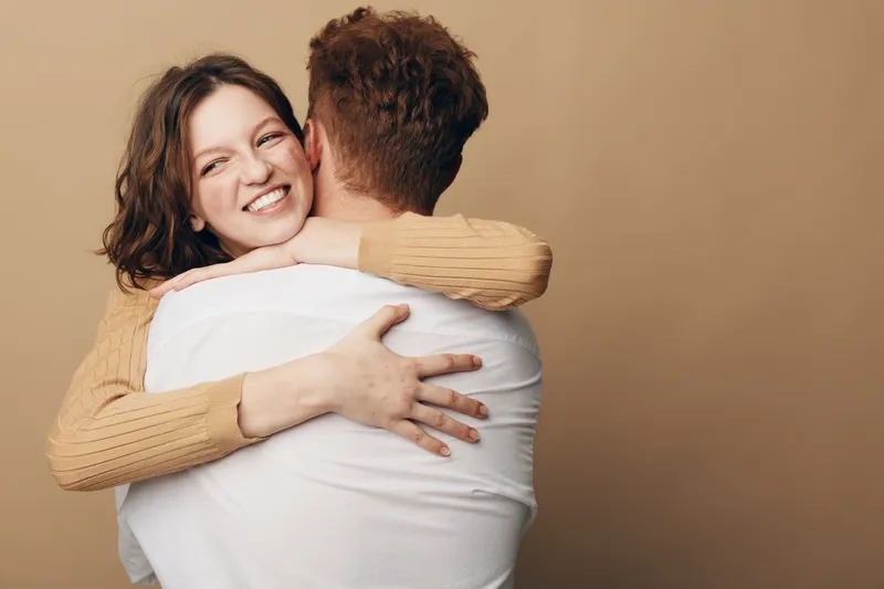 Happy woman with curly hair hugs man wearing white shirt, smiling warmly in beige studio. Concept of love, affection, relationship, intimacy, and emotional connection between couple.