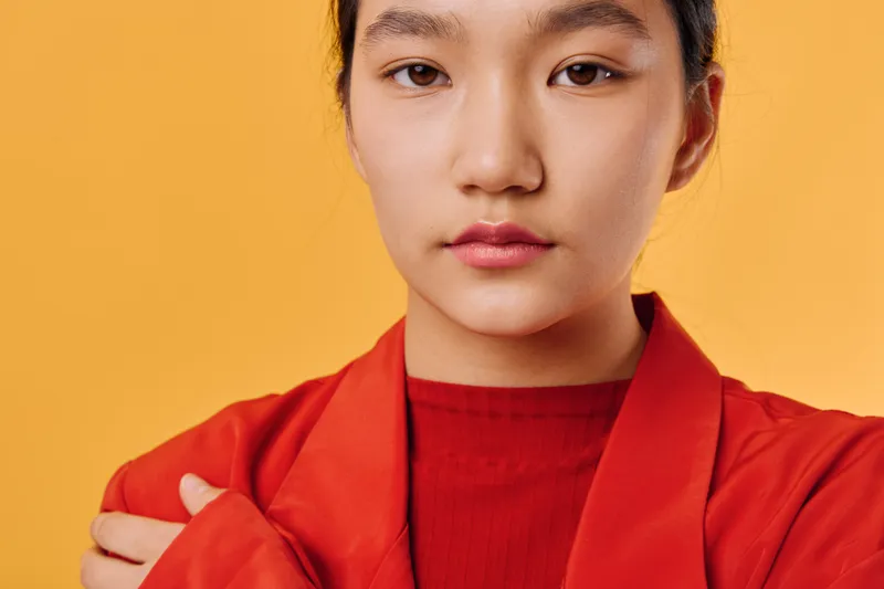 Woman portrait with natural skin and serious expression wearing red jacket against orange background. Closeup face with smooth makeup and minimal accessories in studio light.