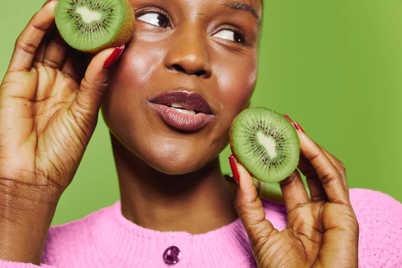 Close-up of a smiling dark skinned African woman holding fresh kiwi slices near her face. Concept for healthy eating, natural beauty, skincare, and wellness on green background.