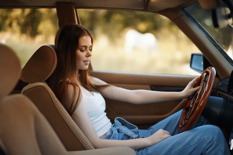 A young woman driving a vintage car, showcasing confidence and style in a casual outfit with a serene expression against a nature backdrop.