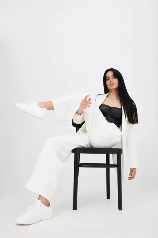 confident woman dressed in white suit and black top posing with relaxed attitude on black chair against plain white background, stylish modern fashion concept